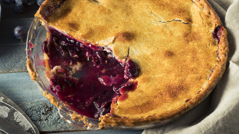 Grape pie in a glass serving dish atop a green slat table missing a large chunk