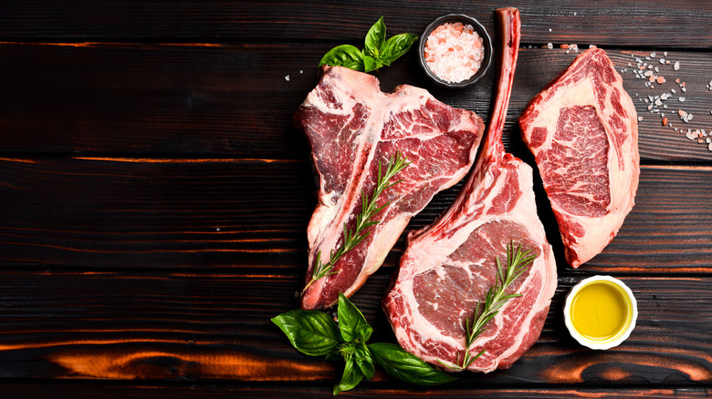 A raw tomahawk, ribeye, and T-bone steak next to seasonings