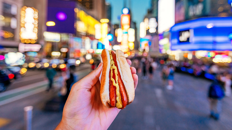 Close up of a hand holding a hot dog against a blurry background of a busy city street