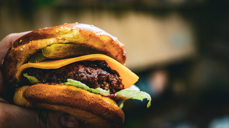 A hand holds big beefy burger with pickles, cheese, and lettuce