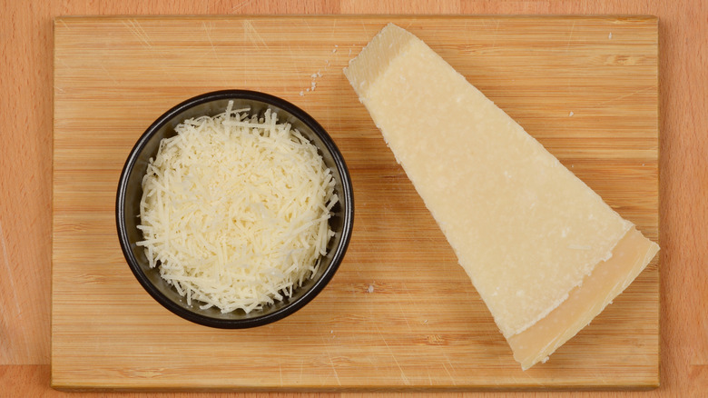 A wedge of parmesan beside a black bowl of grated cheese atop a wooden board
