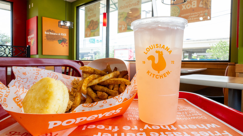 A tray of Popeyes food sits on a tray in a restaurant, including a drink a biscuit, fries, and chicken