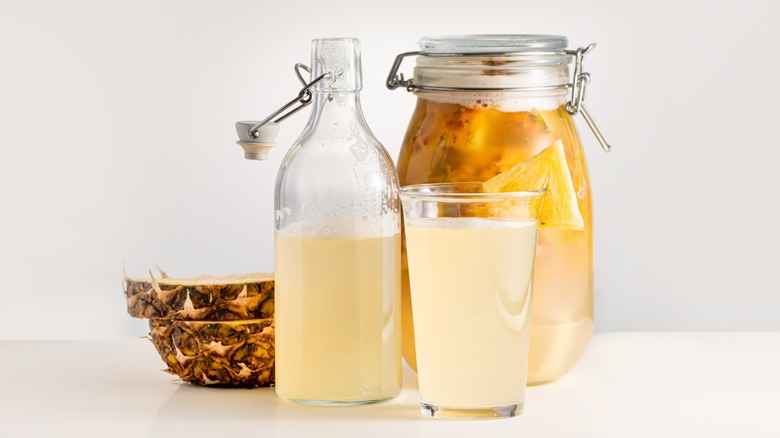 Tepache in a bottle and glass, as well as fermenting with fruit, all next to a cut pineapple