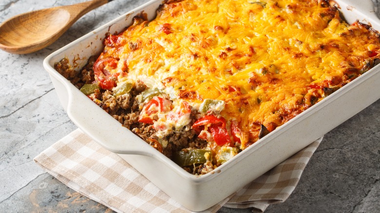 John Wayne Casserole sits in a white dish on a gingham towel with a wooden spoon next to it