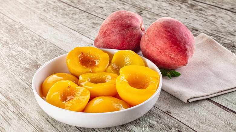 Canned peach halves in a bowl next to fresh peaches.