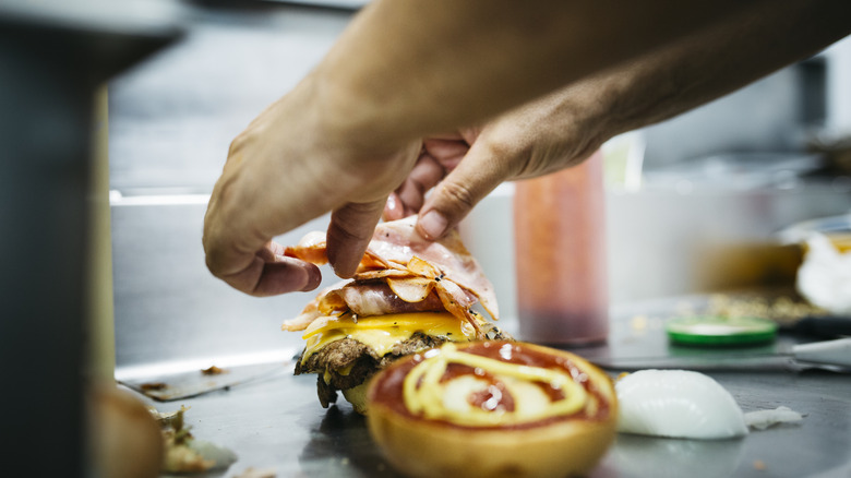 A chef places bacon on top of a cheeseburger