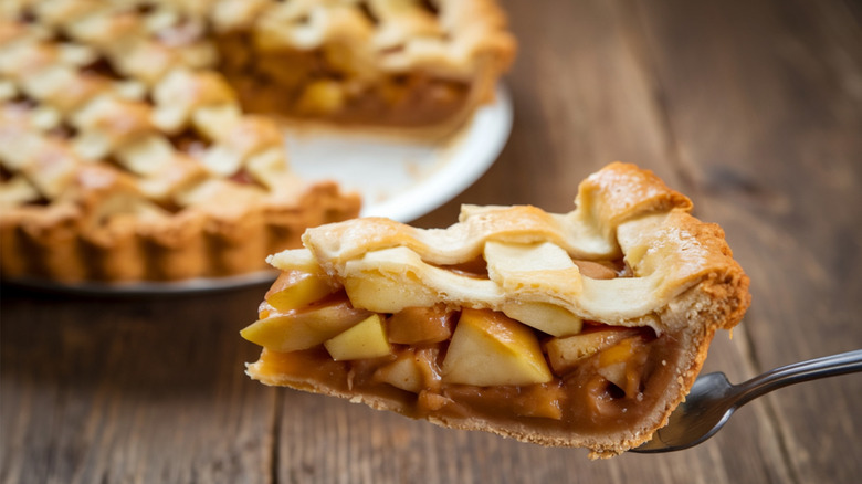 Slice of apple pie held on a fork with the pie it was sliced from in the background on a wooden surface