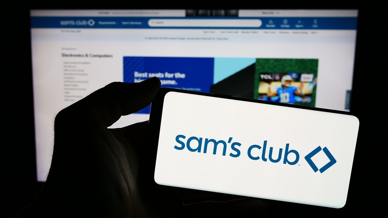 The Sam's Club app is visible on a phone being held up before a computer with the Sam's Club website