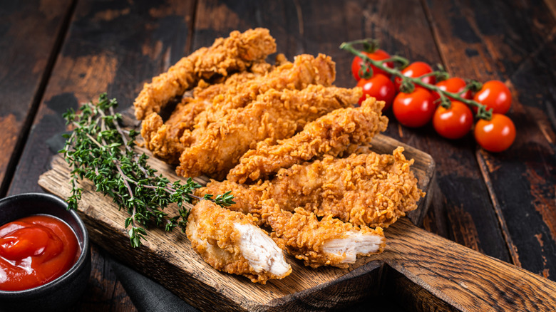Several fried chicken strips on a wooden cutting board with cherry tomatoes, ketchup, and fresh thyme around it