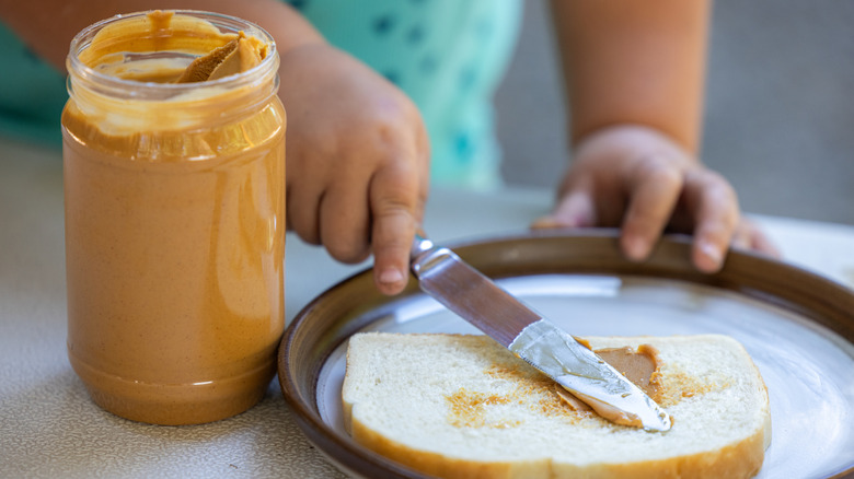 A child's hand spreads peanut butter on a piece of white bread