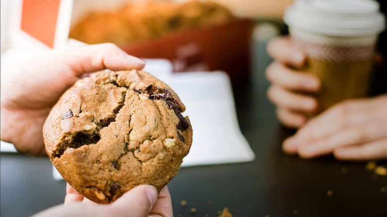 A person holding a chocolate chunk cookie and a person holding a coffee in a to-go cup.