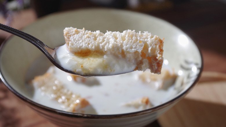 Someone holds up a spoonful of milk toast, a bowl of the breakfast out of focus in the background
