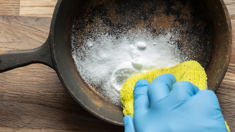 Person with glove using sponge and baking soda to clean rust off cast iron pan.