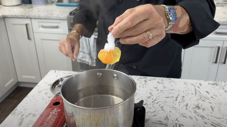 Hand holding egg in saran wrap cling film over pot of water