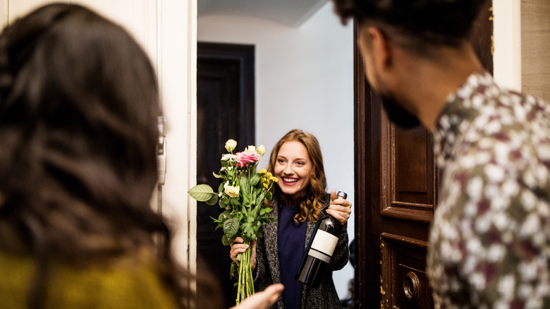 Woman with wine bottle and bouquet of flowers welcomed by friends at door