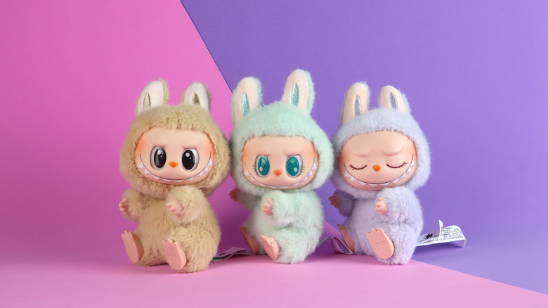 Three Labubu monsters sit against a pink and purple background